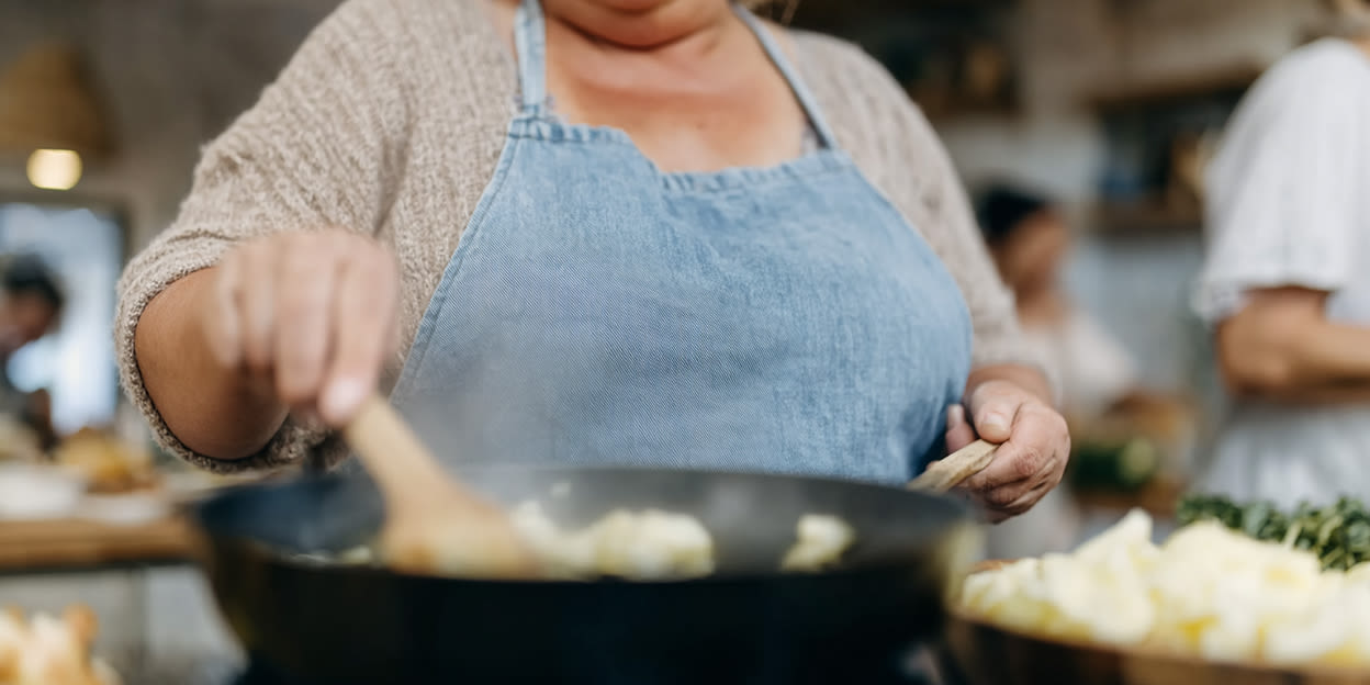 Kvinna med blått förkläde står och rör runt i en stekpanna på spisen. Bilden är delvis skapad med AI.