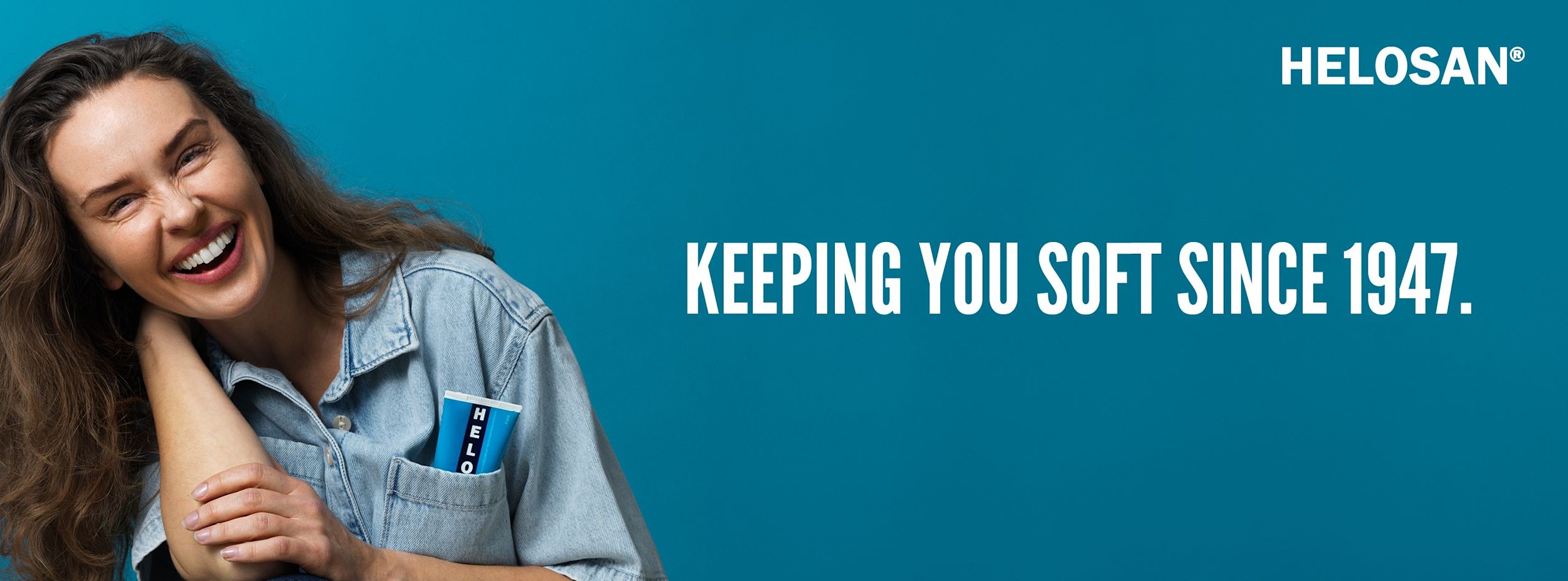 Leende kvinna i jeansskjorta håller armen om sig själv, tub Helosan i bröstfickan, text: "KEEPING YOU SOFT SINCE 1947."