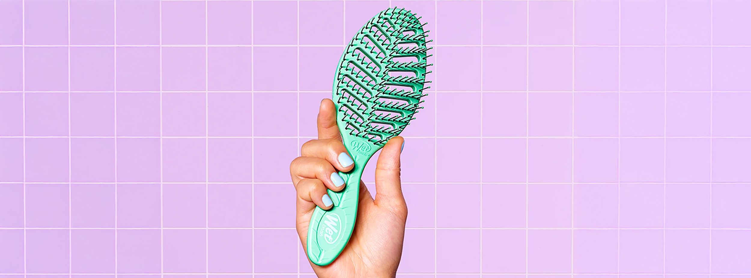 Hand håller en grön Wet Brush-hårborste framför ett lila kaklat väggmönster.
