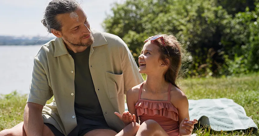 Pappa och dotter sitter utomhus på en filt i solen. Dottern har smetat in solskydd på pappans panna och skrattar.