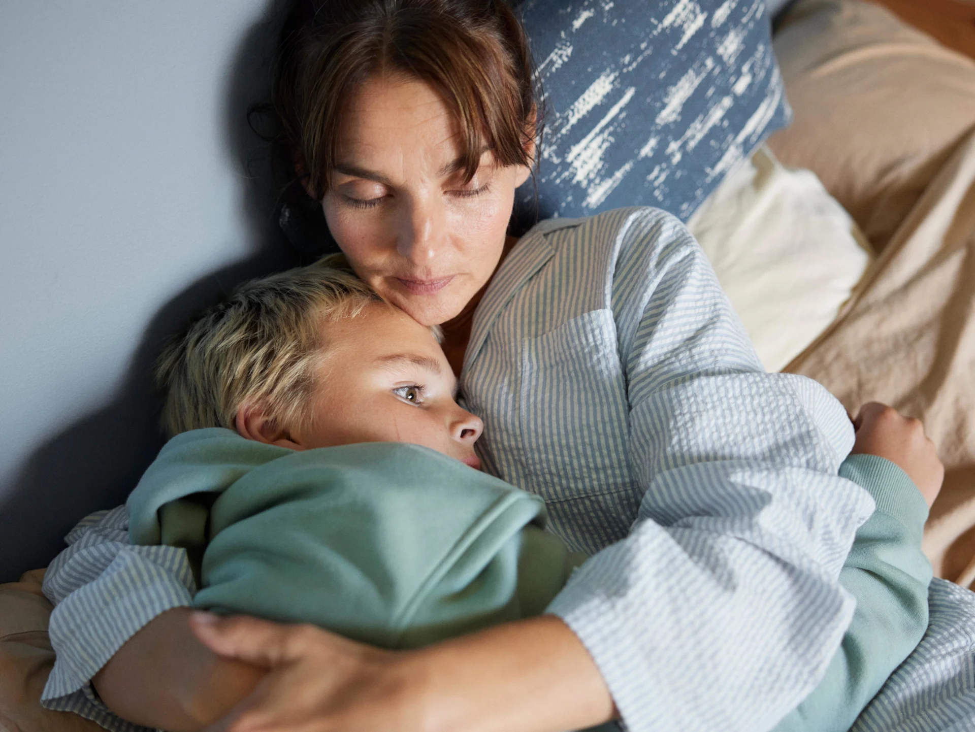 Mamma och son ligger i sängen och vilar och kramas. 