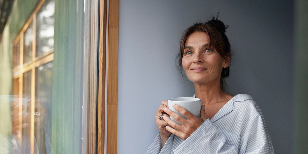 Kvinna står vid ett fönster med en kopp i handen. Hon kollar ovanför kameran.