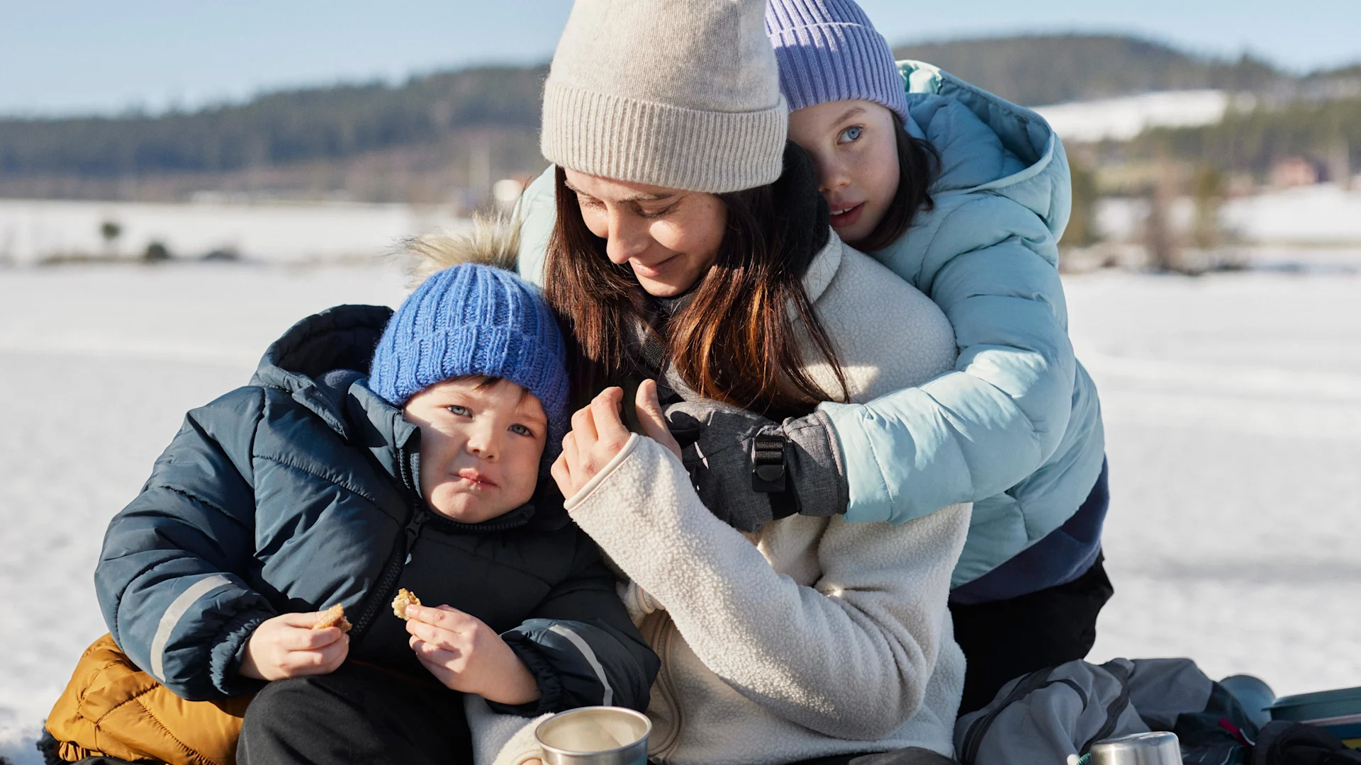 Mamma sitter med två barn utomhus i snön, iklädda mössor och varma jackor och fikar tillsammans.