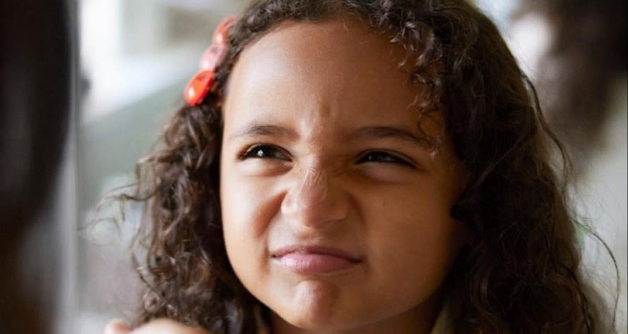 Siluetten av en förälder syns snett framför ett barn med mörkt, långt och lockigt hår med ett rött hårspänne. Barnet har en trumpen min och ser inte helt nöjd ut. 