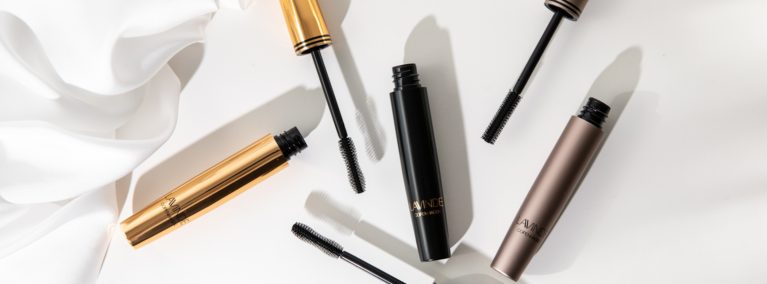 Tre öppna mascaror i guld, svart och brons med texten "LAVINDE COPENHAGEN" ligger på ett vitt underlag bredvid vit tyg.