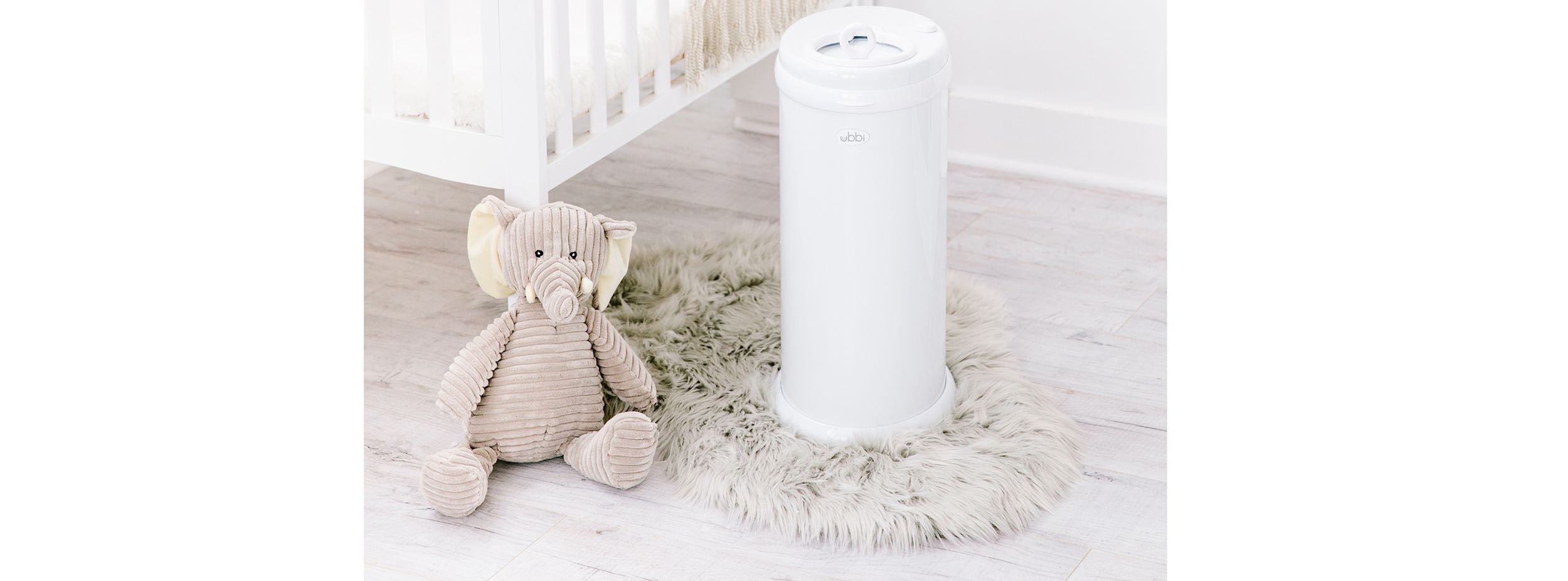 En beige elefantmjukisdjur sitter på en lurvig matta bredvid en vit Ubbi blöjhink intill en spjälsäng i ett ljust barnrum.