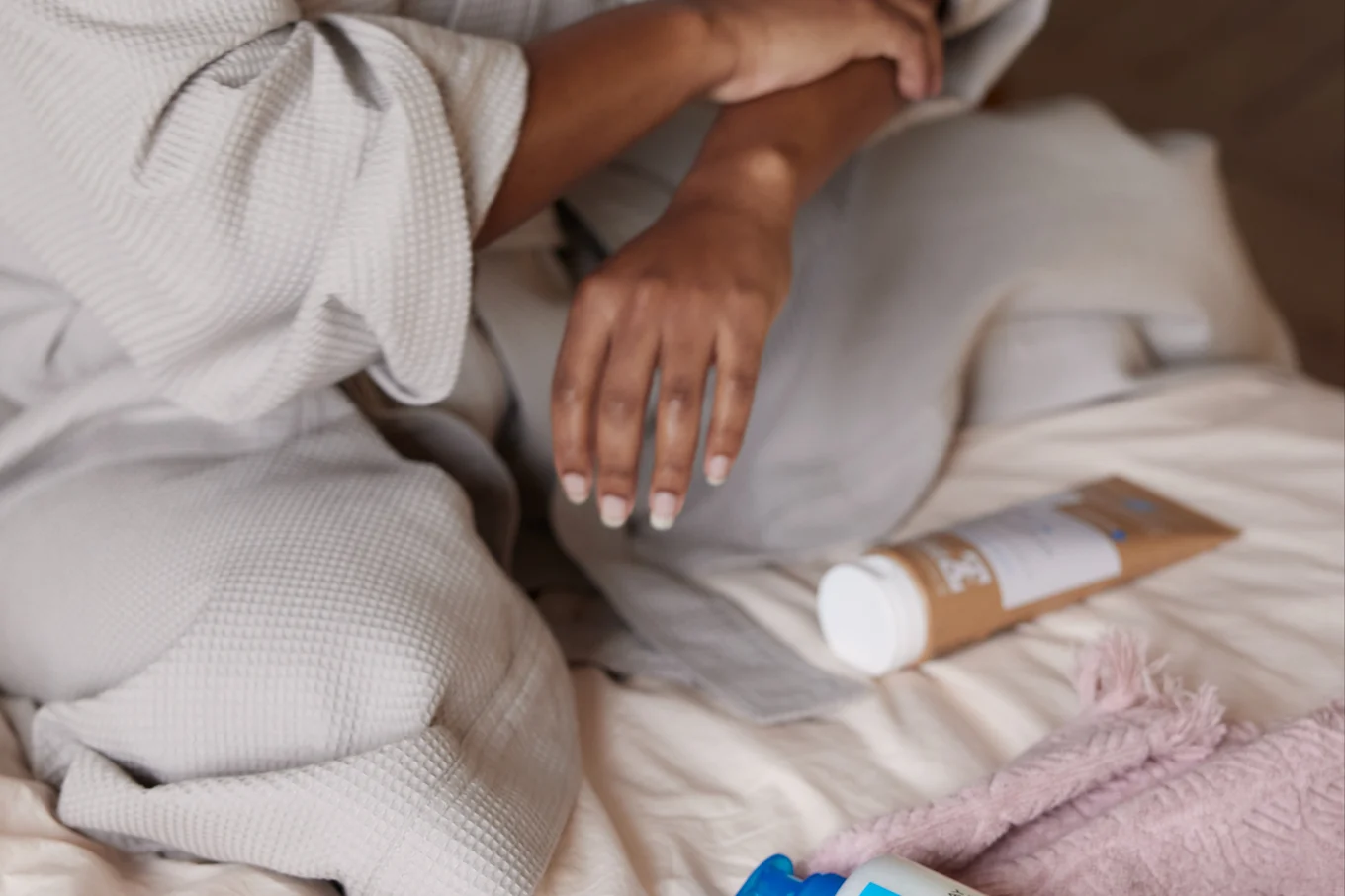Kvinna sitter i morgonrock i en säng och smörjer in sin arm med hudkräm som återfuktar huden