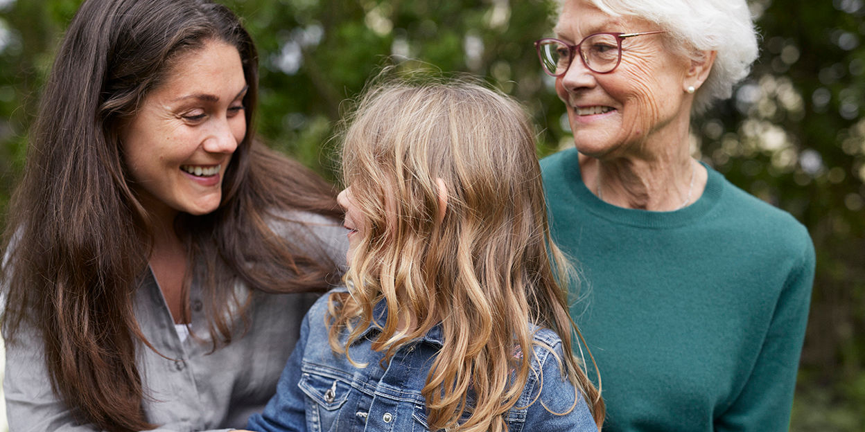 En mormor, en mamma och en liten flicka sitter utomhus och blickar kärleksfullt mot varandra.