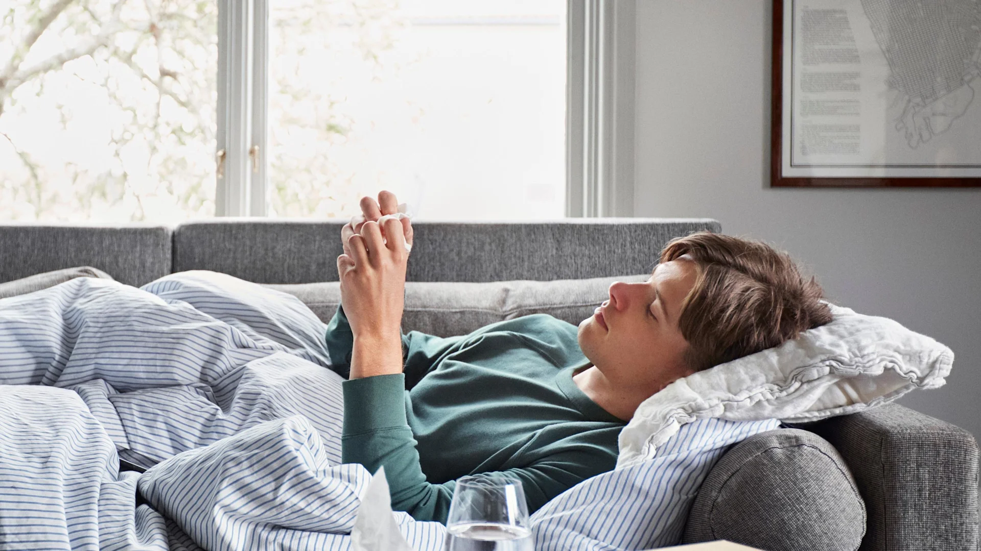 Man ligger nedbäddad i soffan med täcke och kuddar, med näsdukar runtomkring sig.