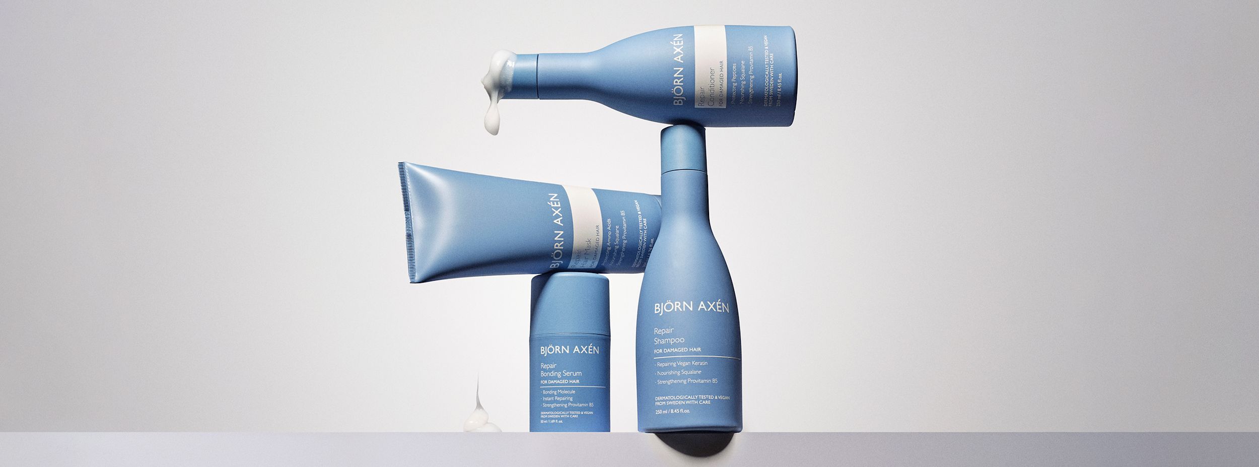 Tre blå Björn Axén hårvårdsprodukter står staplade, med schampo, balsam och inpackning, mot ljus bakgrund. Kräm rinner från balsamflaskan.