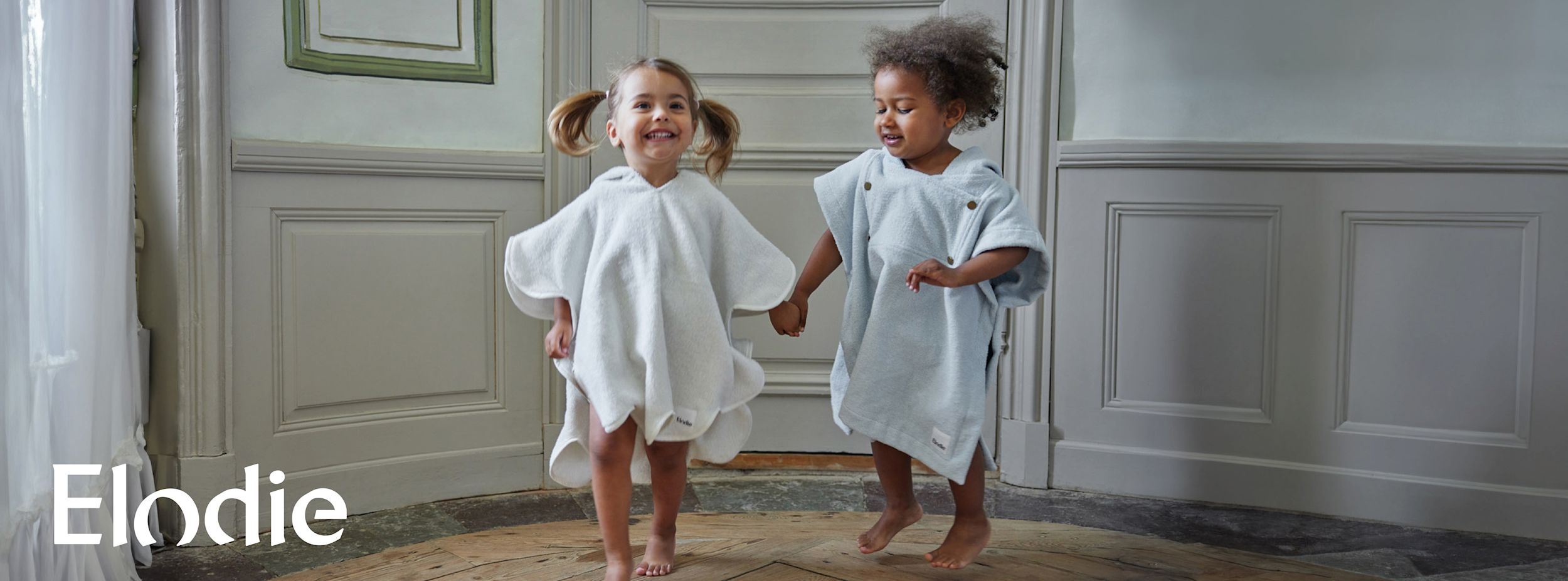 Två små barn i ponchos håller varandra i handen och hoppar glatt på ett trägolv i ett ljust rum. Text: Elodie.
