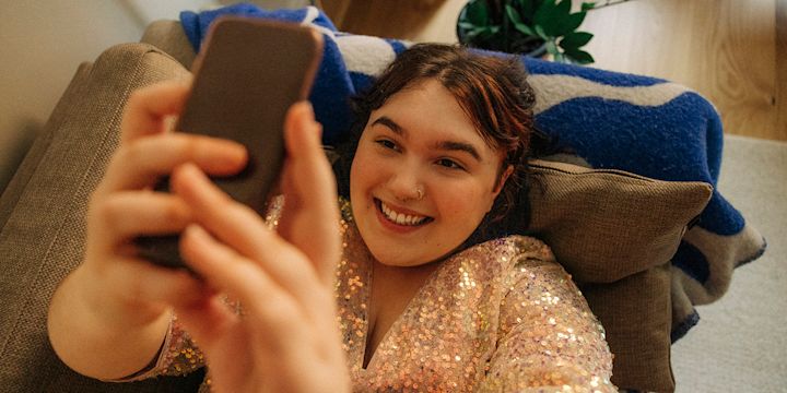 Glad tjej med glittrig klänning ligger i soffan och tittar i sin mobil.