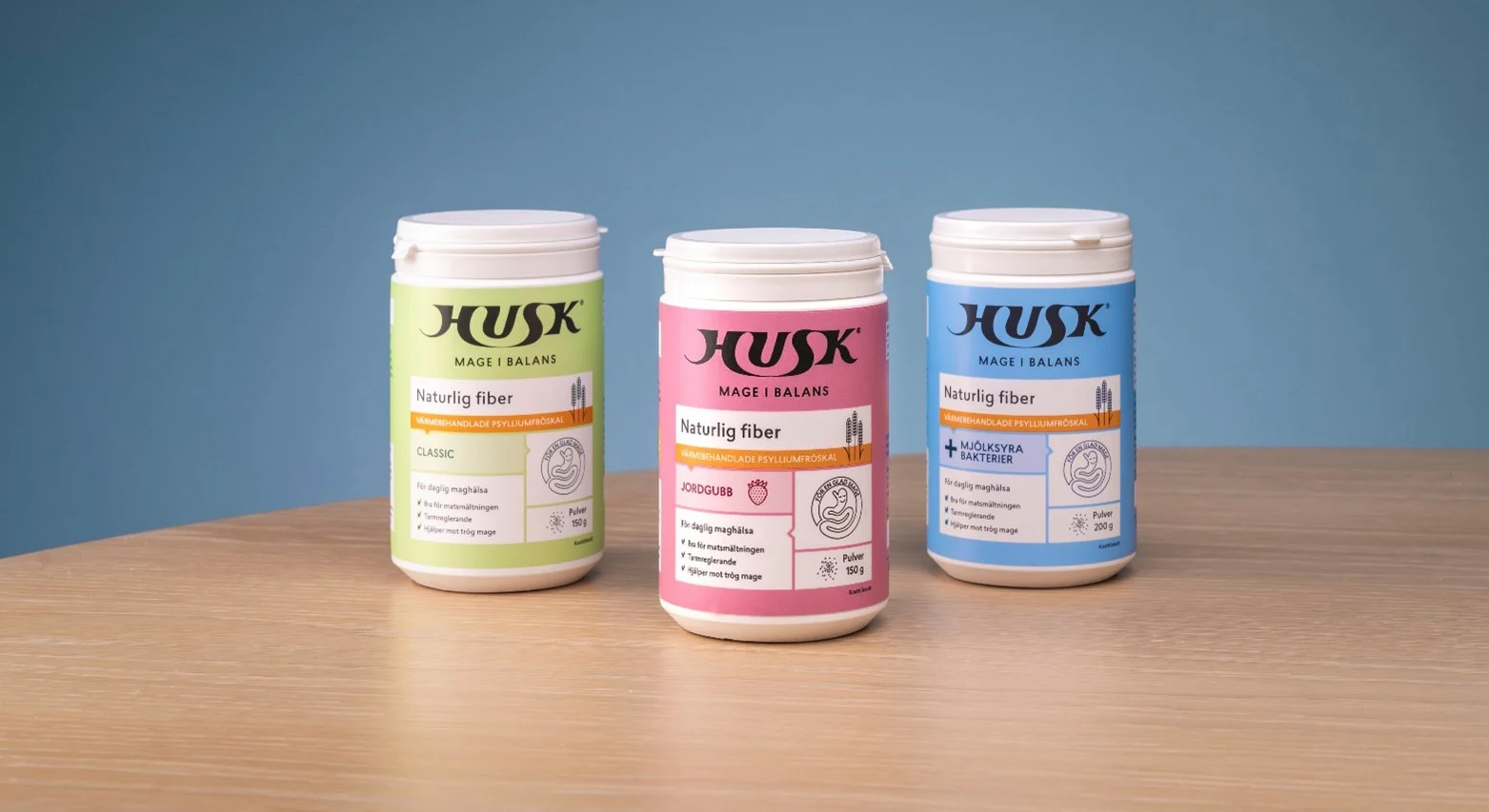 Tre behållare med Husk naturfiber i grönt, rosa och blått, uppsatt på ett träbord mot en blå bakgrund.