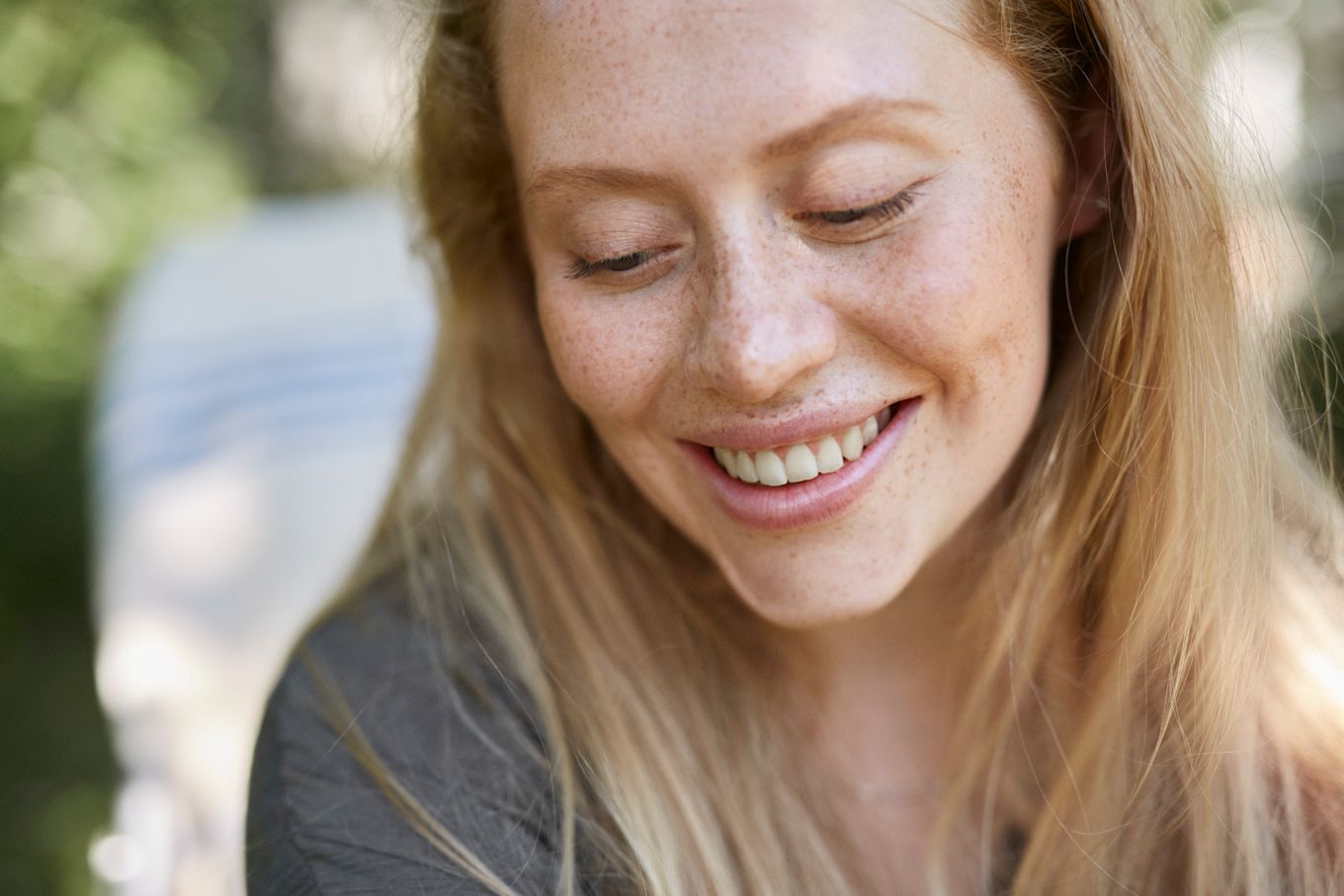 Ung kvinna med fräknigt ansikte sitter utomhus i soligt väder och ler stort.