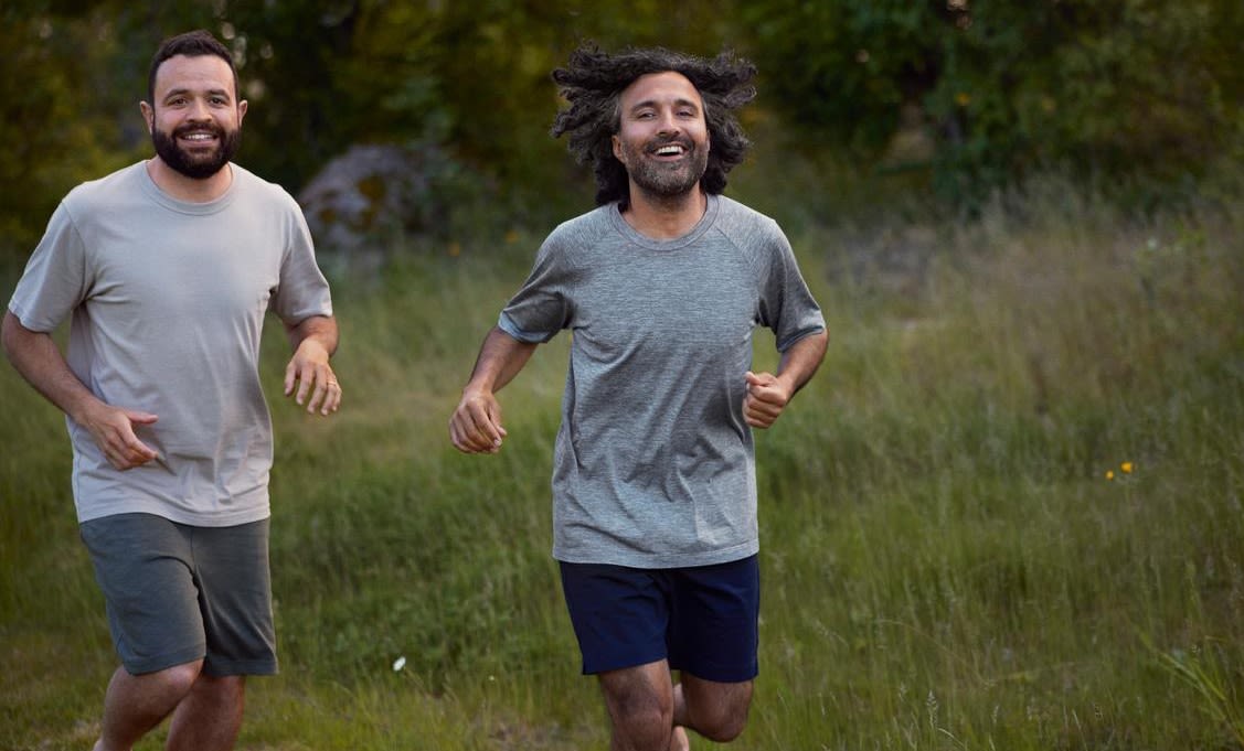 Två glada män med mörkt hår och helskägg är ute och springer tillsammans. De har grå t-shit och mörkgröna respektive mörkblå shorts. De springer ute på öppen gräsmark med skog i nära anslutning.
