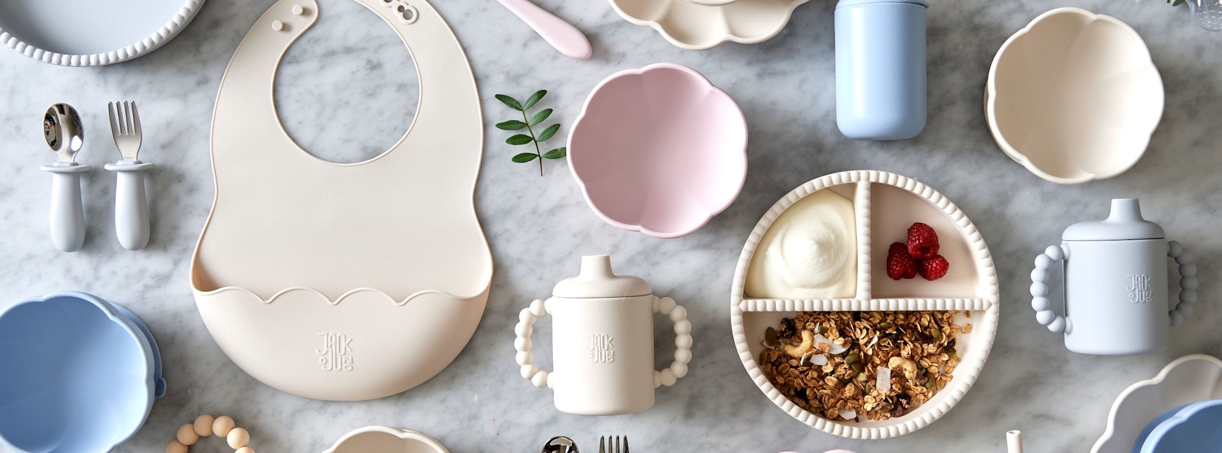 Barnservis i pastellfärger med tallrik, skål, haklapp, pipmugg, bestick och delad tallrik med yoghurt, granola och hallon på marmorbord.