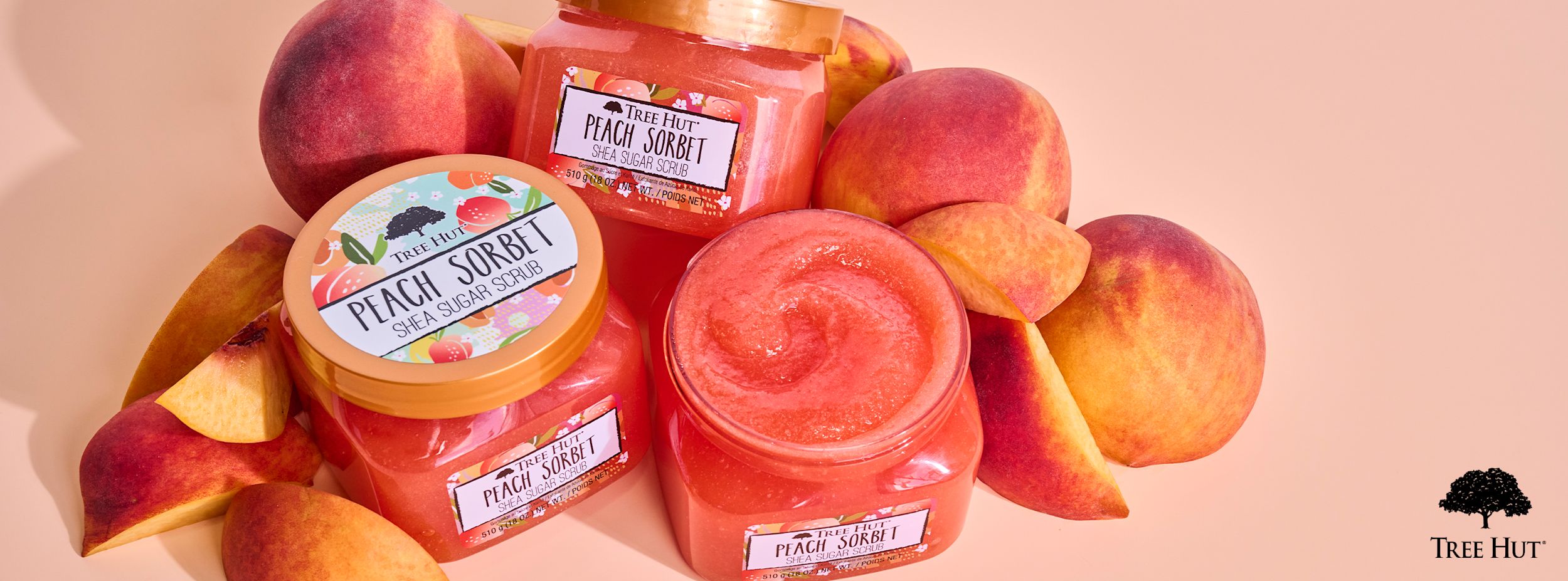 Tre Hut Peach Sorbet Shea Sugar Scrub-burkar står bland färska persikor på en ljus bakgrund, med Tree Hut-logotypen i hörnet.