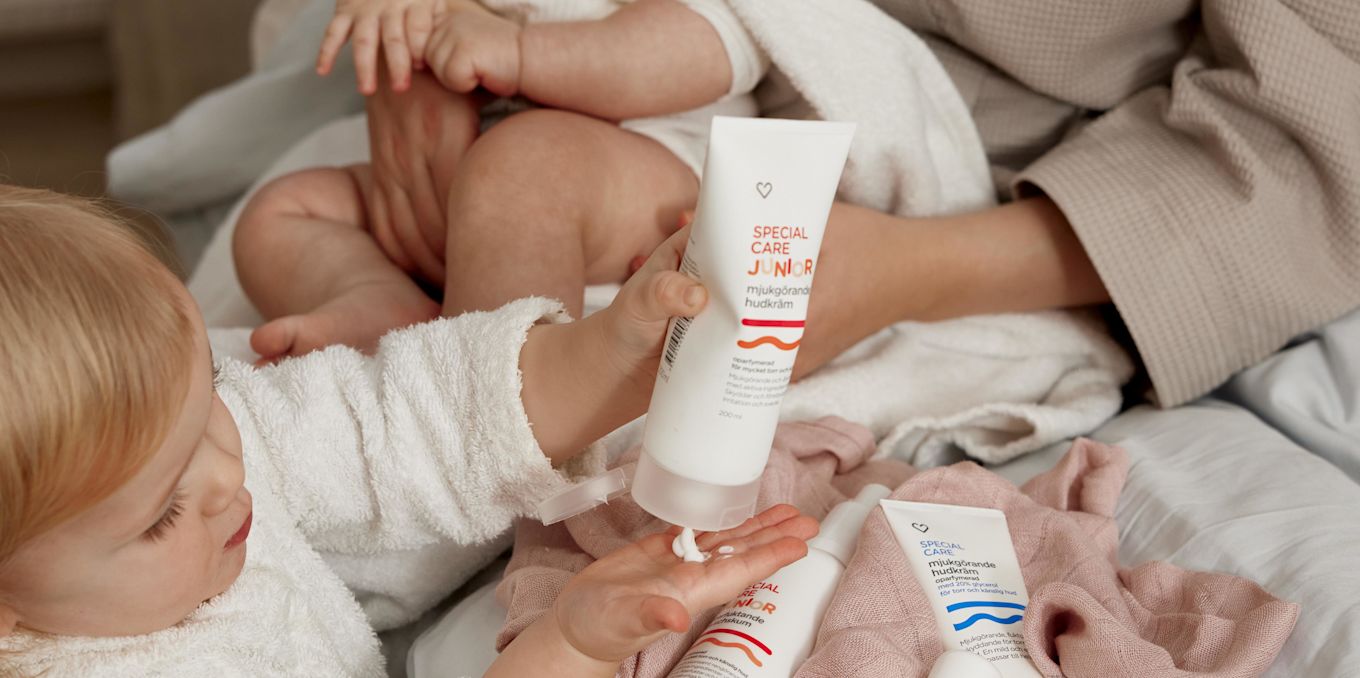 Ett barn i morgonrock pressar ut hudkräm i handen. I bakgrunden syns en mamma med sin bebis i famnen, hon sitter på sängkanten. 