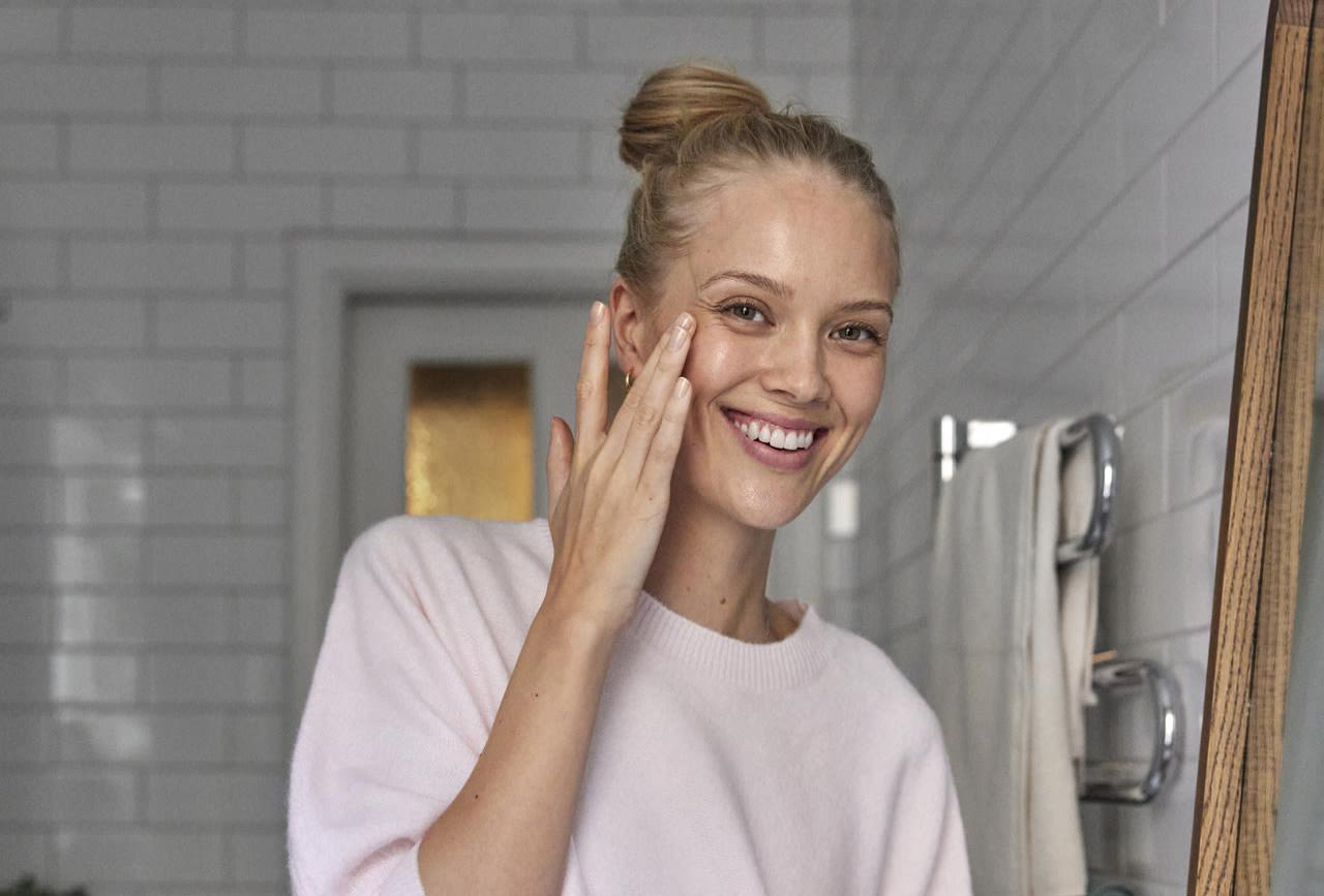 En glad tjej står i badrummet och smörjer in sin ena kind med ena handen. Hon är glad och tittar in i kameran.