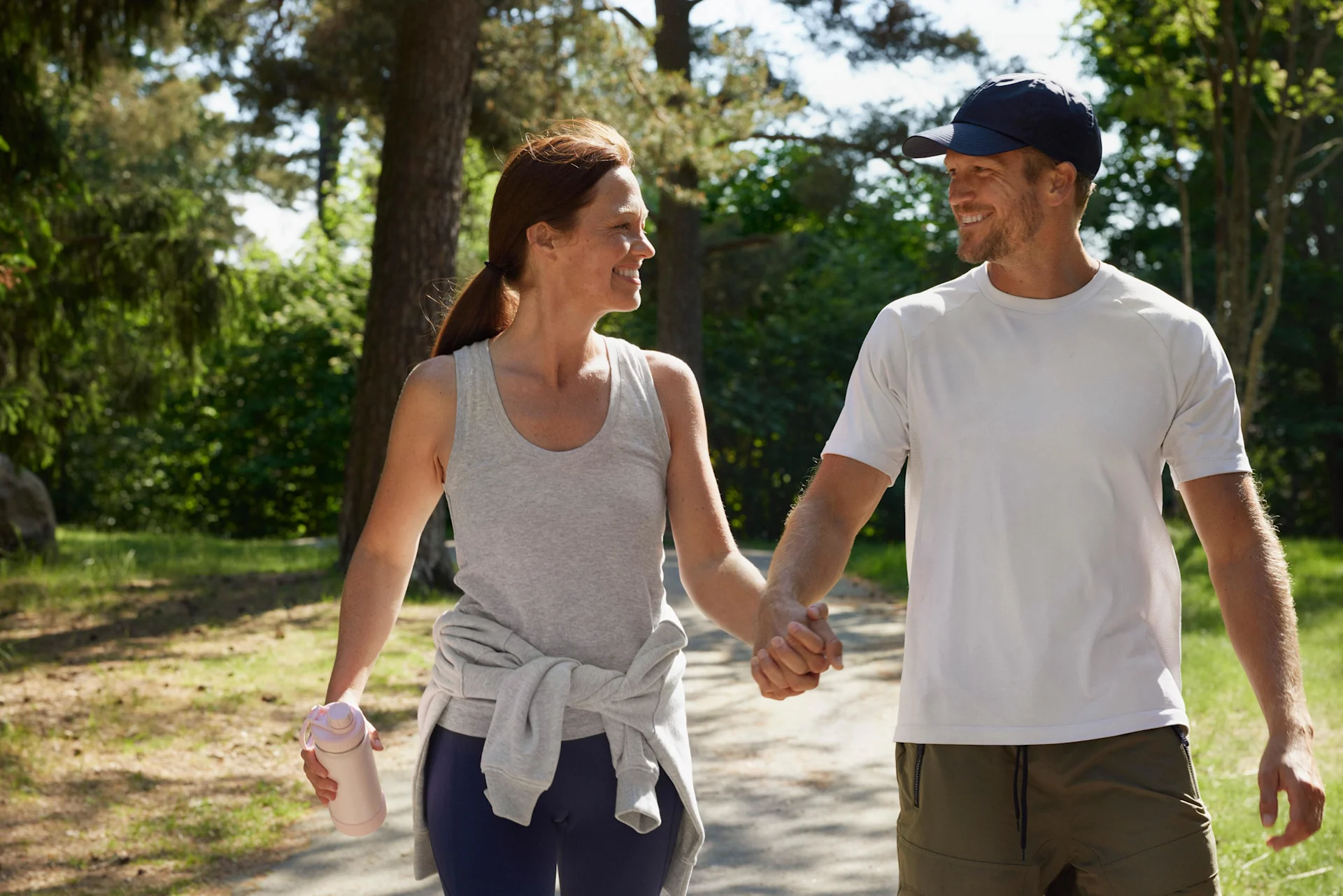 En kvinna och man är ute och går på en promenad i en solig, öppen skog. De håller varandra i handen och tittar på varandra och ler. Kvinnan håller i en vattenflaska och det ser ut som att det är varmt ute. 