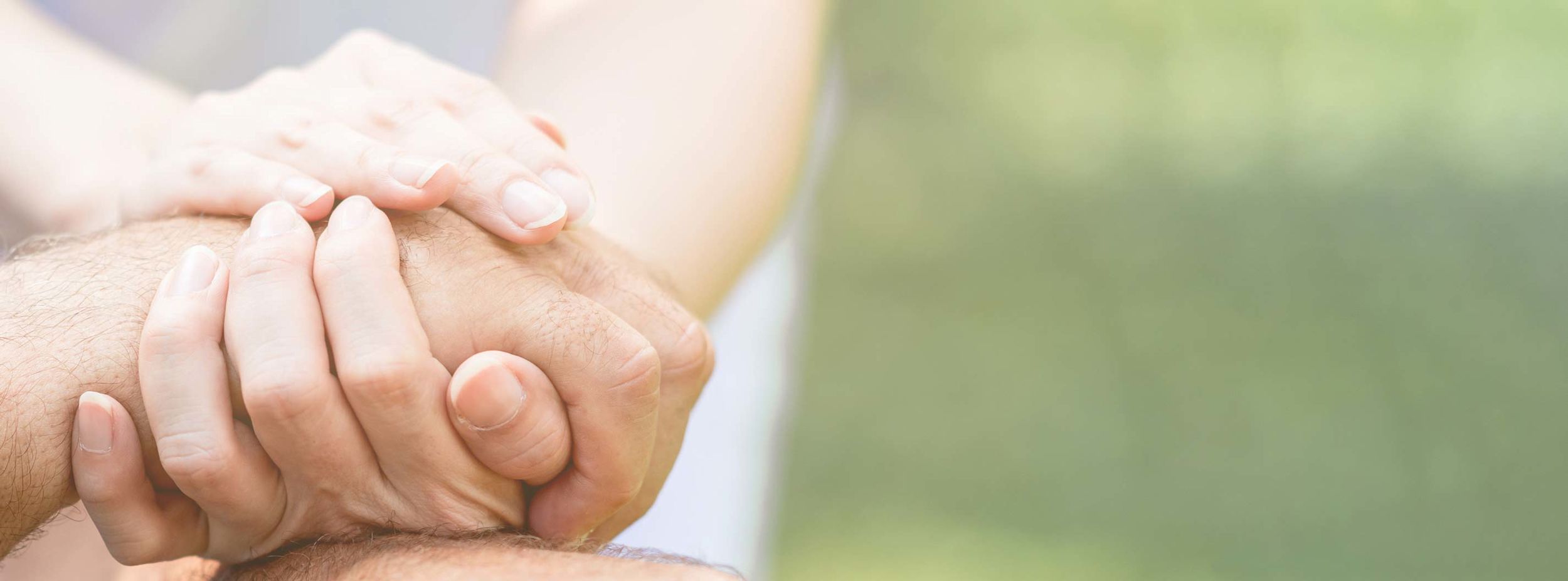 Två händer håller varsamt om en annan hand som en gest av stöd och omtanke, med suddig grön bakgrund.