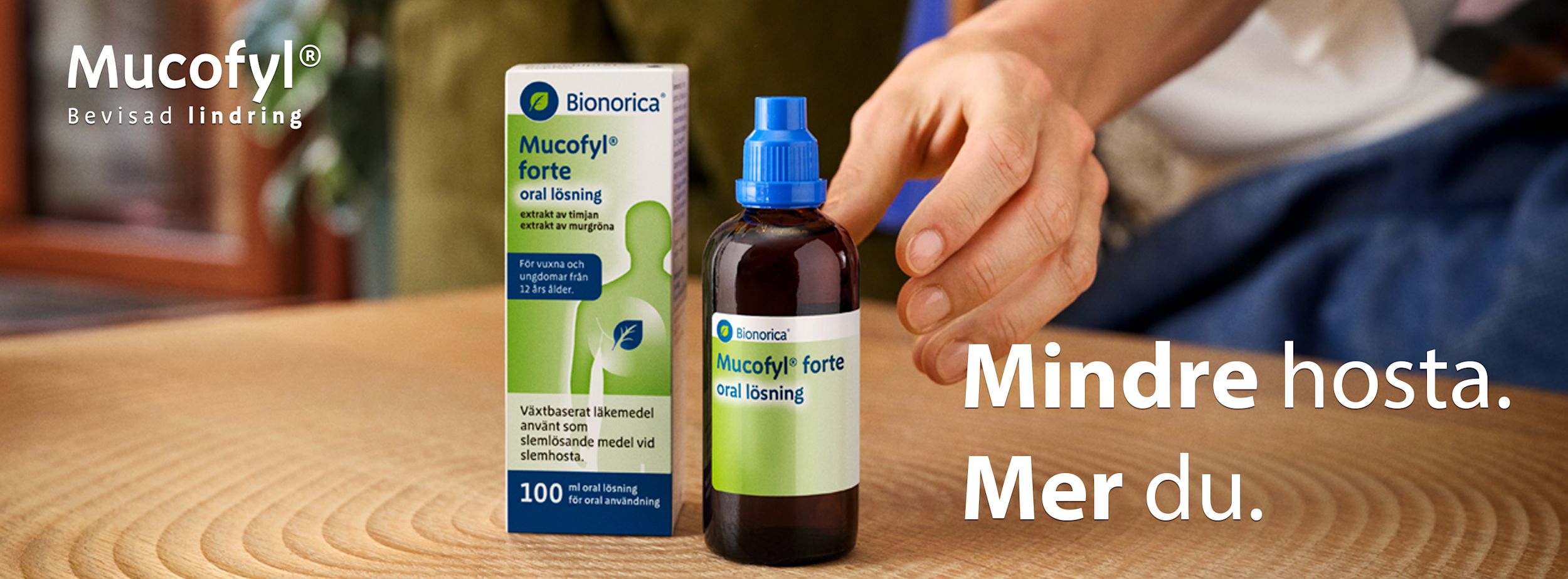 En hand når efter en flaska Mucofyl forte oral lösning bredvid förpackningen på ett bord, text: "Mindre hosta. Mer du.".
