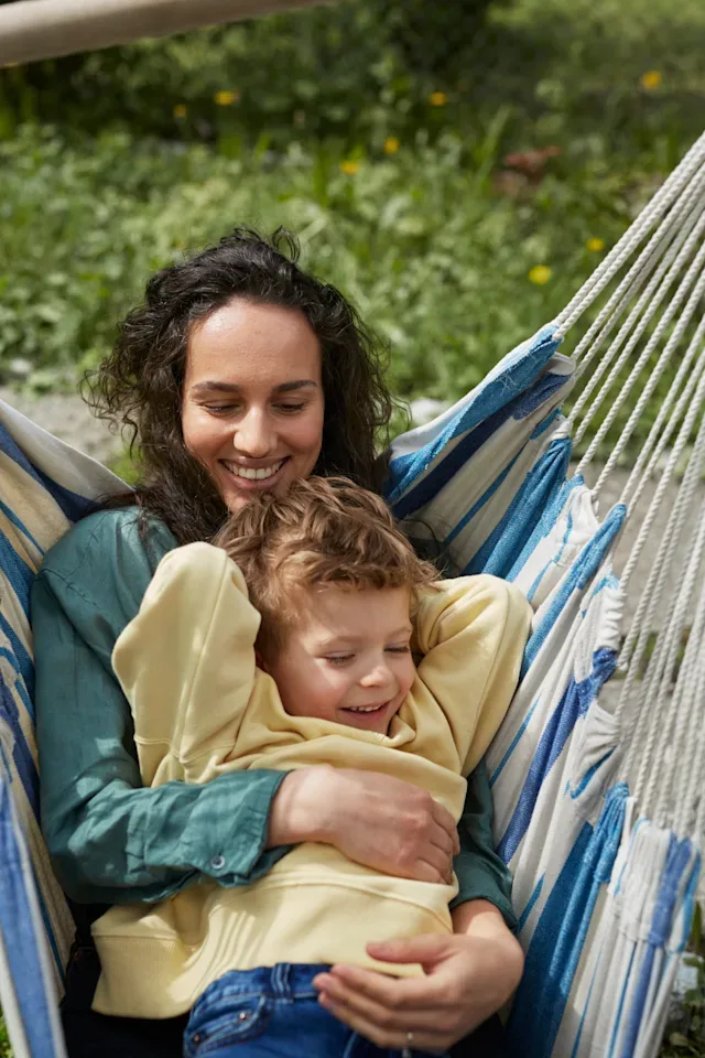 En mamma och son sitter i en hängmatta i en somrig trädgård trädgård. De skrattar och kramas.