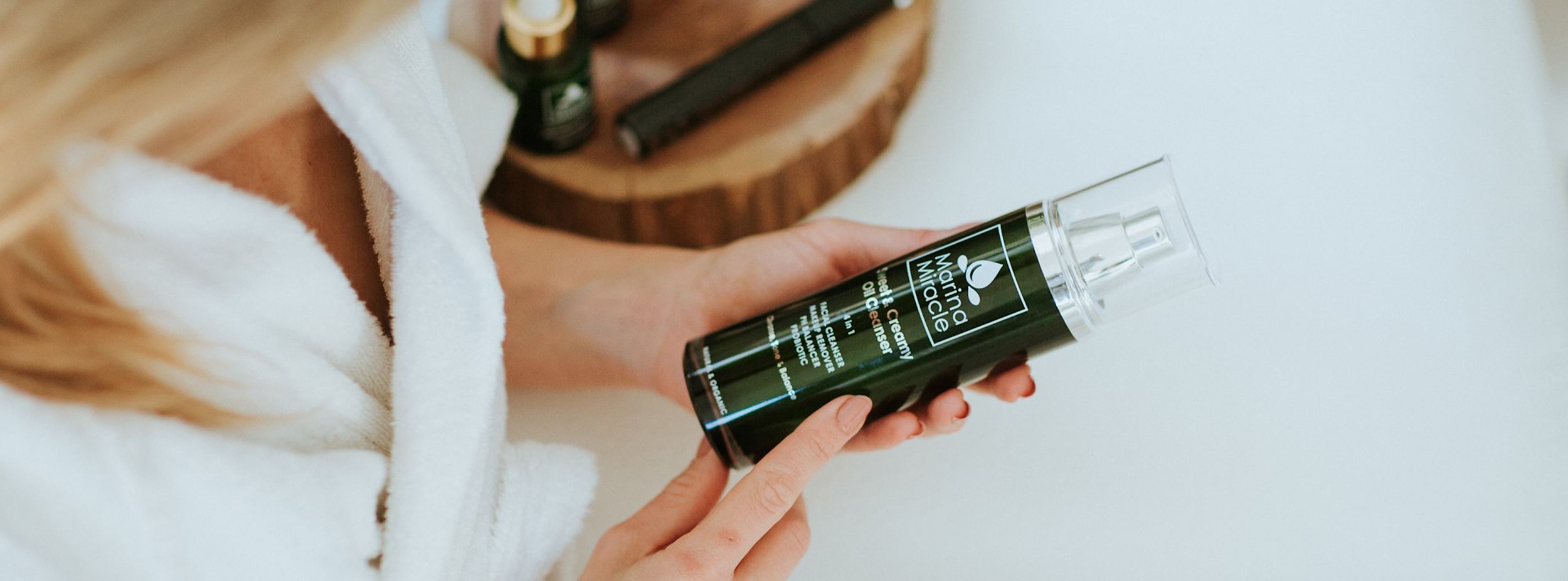Kvinna i vit morgonrock håller en flaska Marina Miracle Sweet & Creamy Oil Cleanser i händerna i ett badrum.