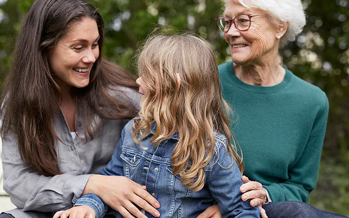 Mamma, mormor och barnbarn sitter tillsammans och ser glada ut.