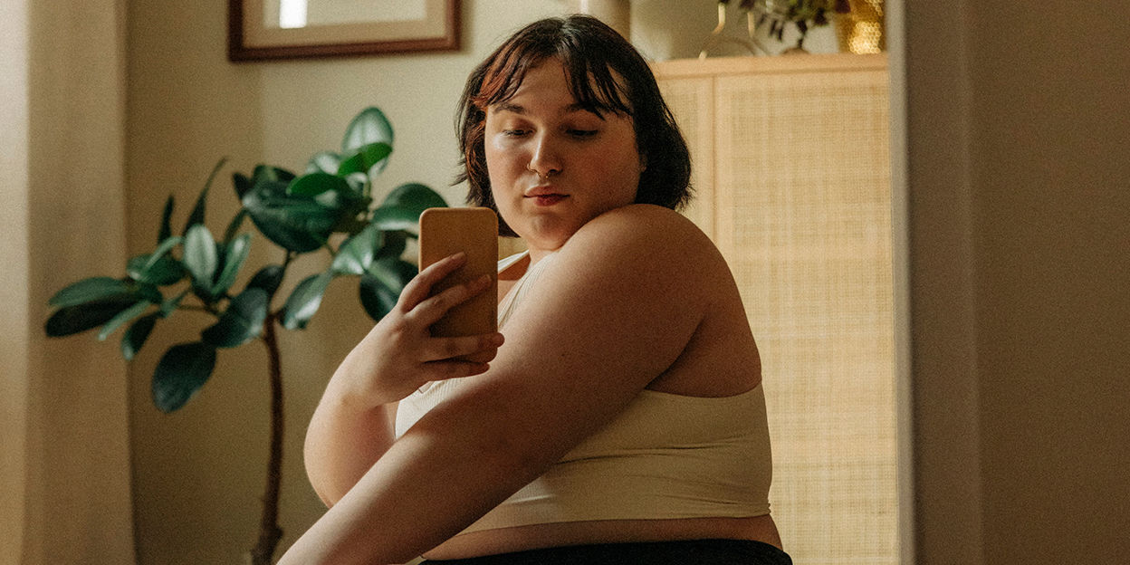 En kjej står i linne och tittar sig i spegeln. Hon håller en mobiltelefon i ena handen och tar en bild av sig själv.