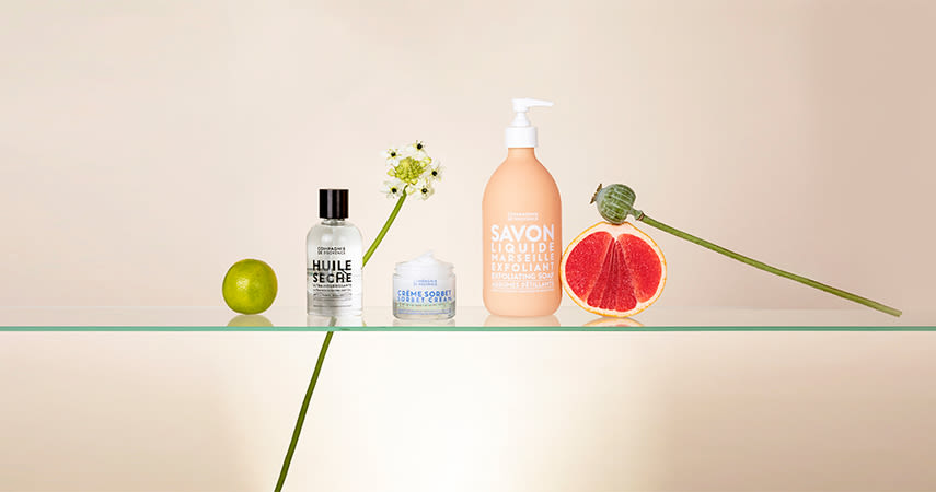 Fyra skönhetsprodukter står på en glasskiva tillsammans med en lime, en blomma och en halv grapefrukt.