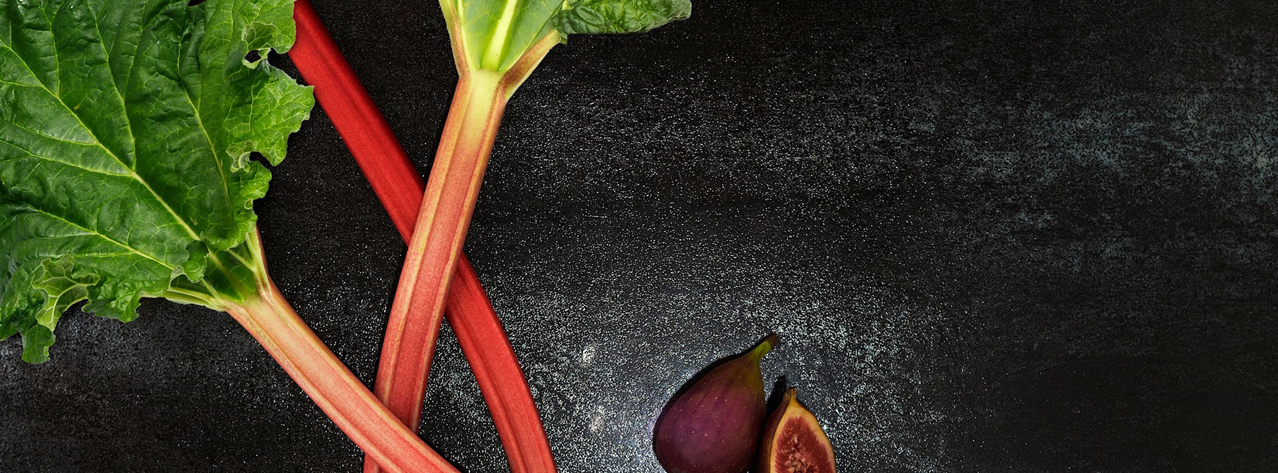 Två rabarberstjälkar med gröna blad ligger bredvid ett helt och ett halvt fikon på en mörk yta.
