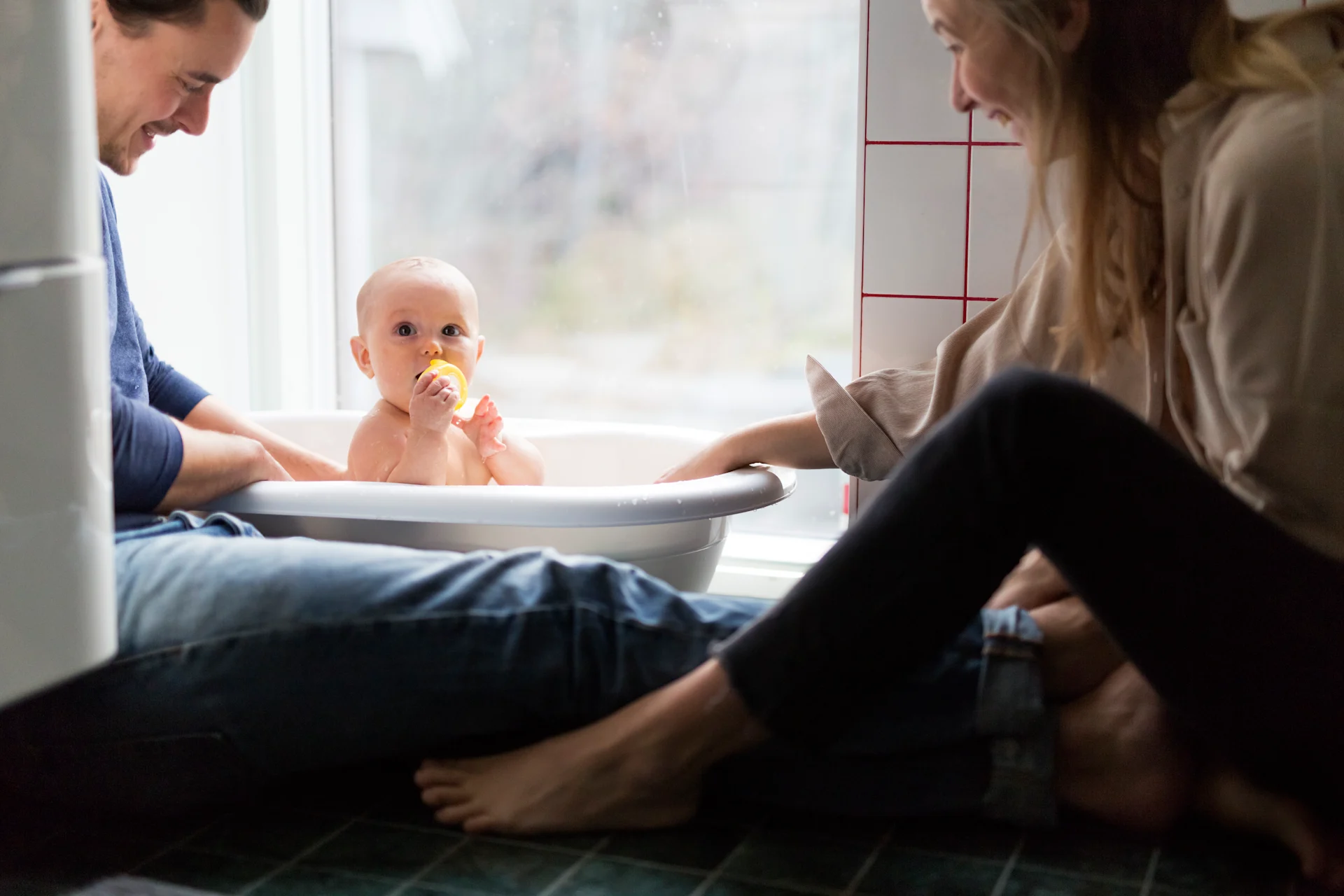 Bebis blir badad i balja av mamma och pappa, leker med en gummianka.