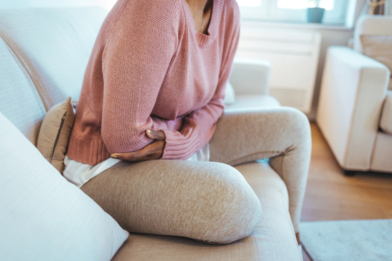 Kvinna sitter och håller om sin mage i soffan hemma, iklädd mjukiskläder.