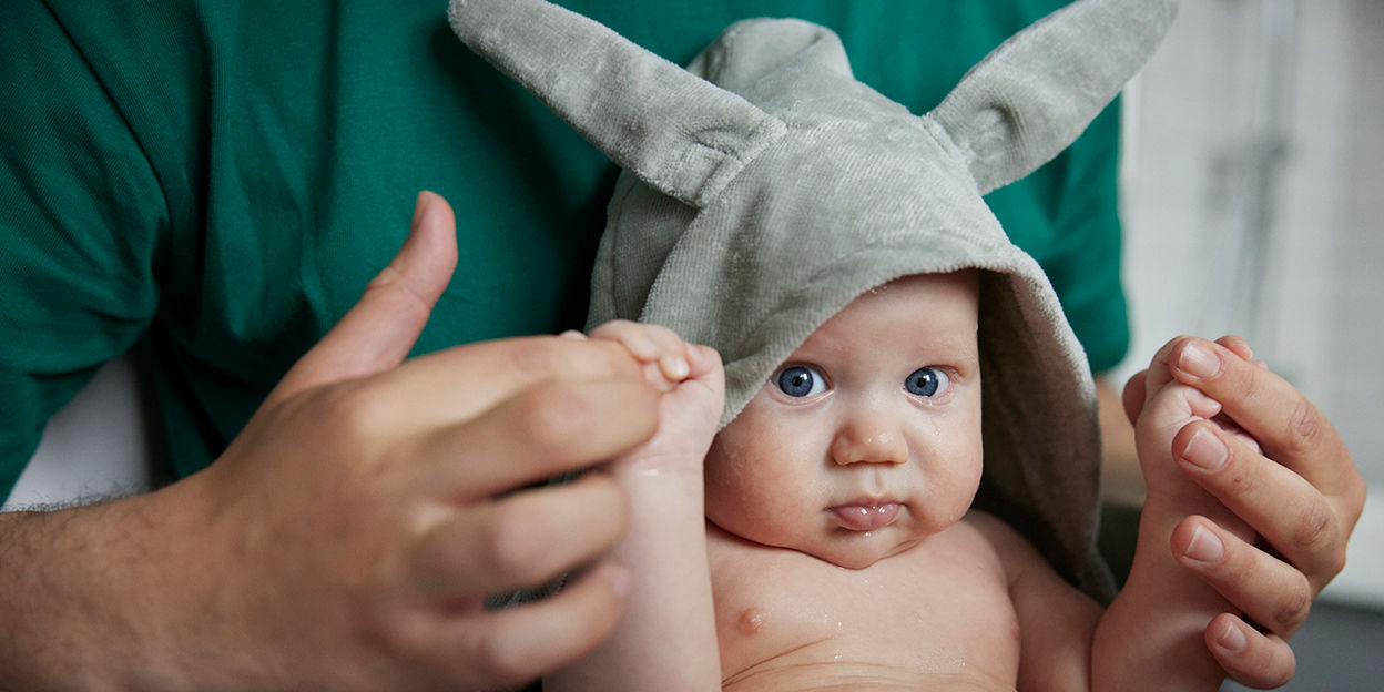Bebis med luva sitter i vuxen persons famn och kollar in i kameran. 