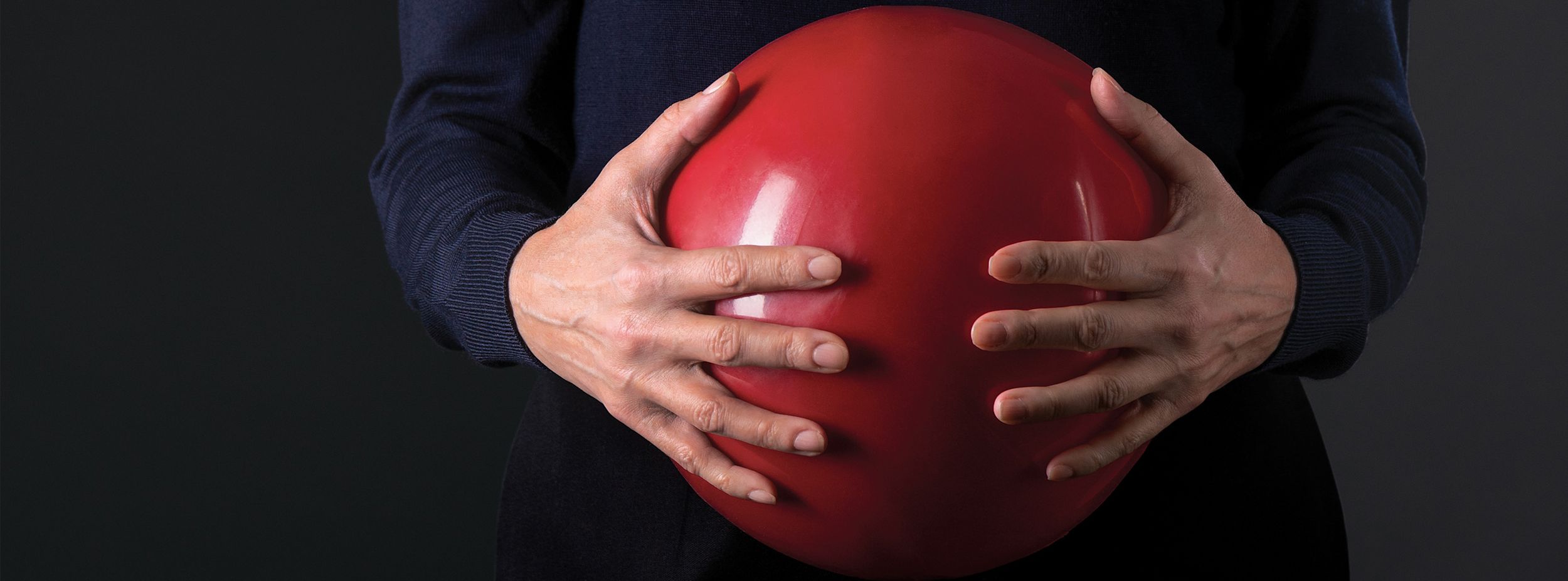Person håller en stor röd boll med båda händerna mot magen, klädd i mörk tröja, mot mörk bakgrund.
