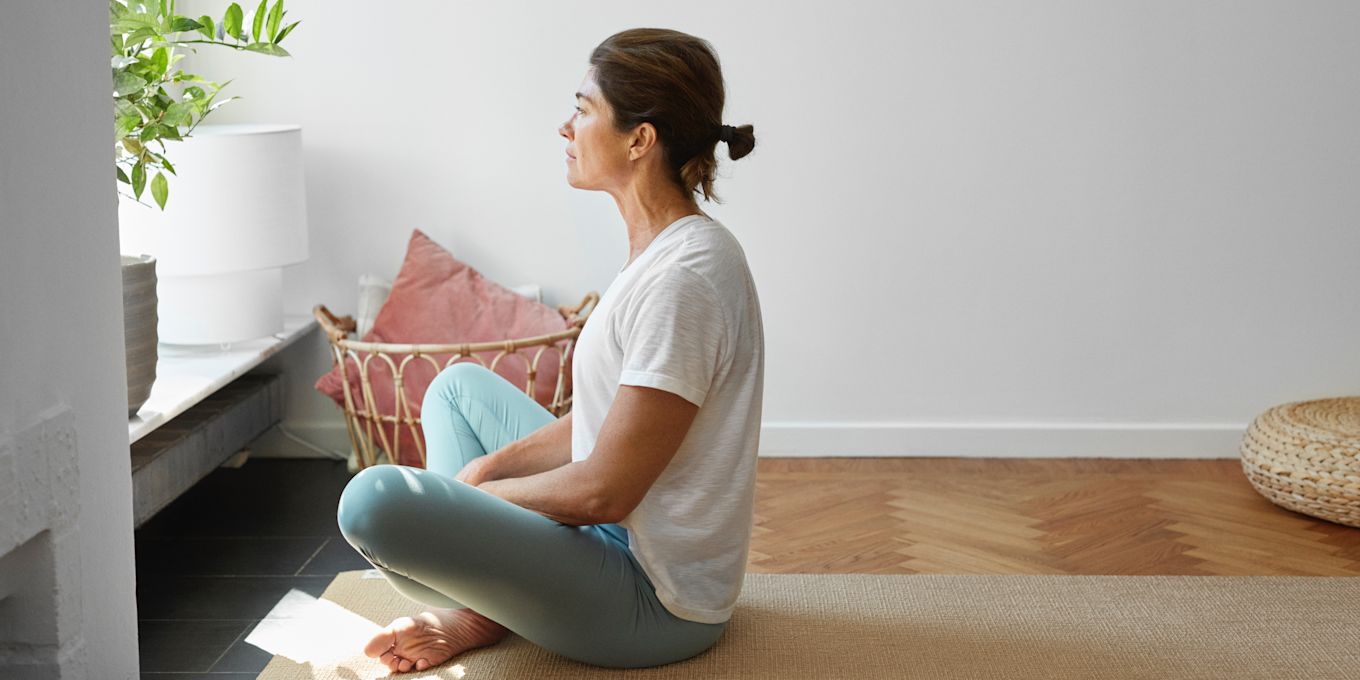 Kvinna sitter på en yogamatta och tittar ut genom fönstret.