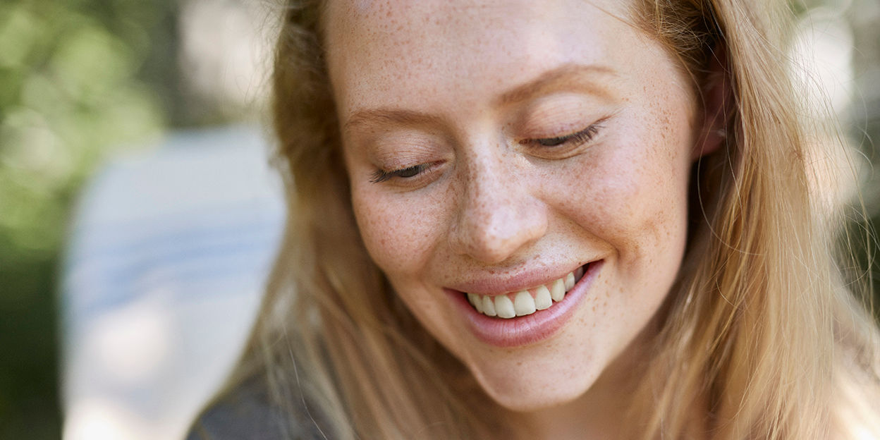 Kvinna ler och kollar snett nedåt, fokus är på hennes ansikte. Hon är utomhus.