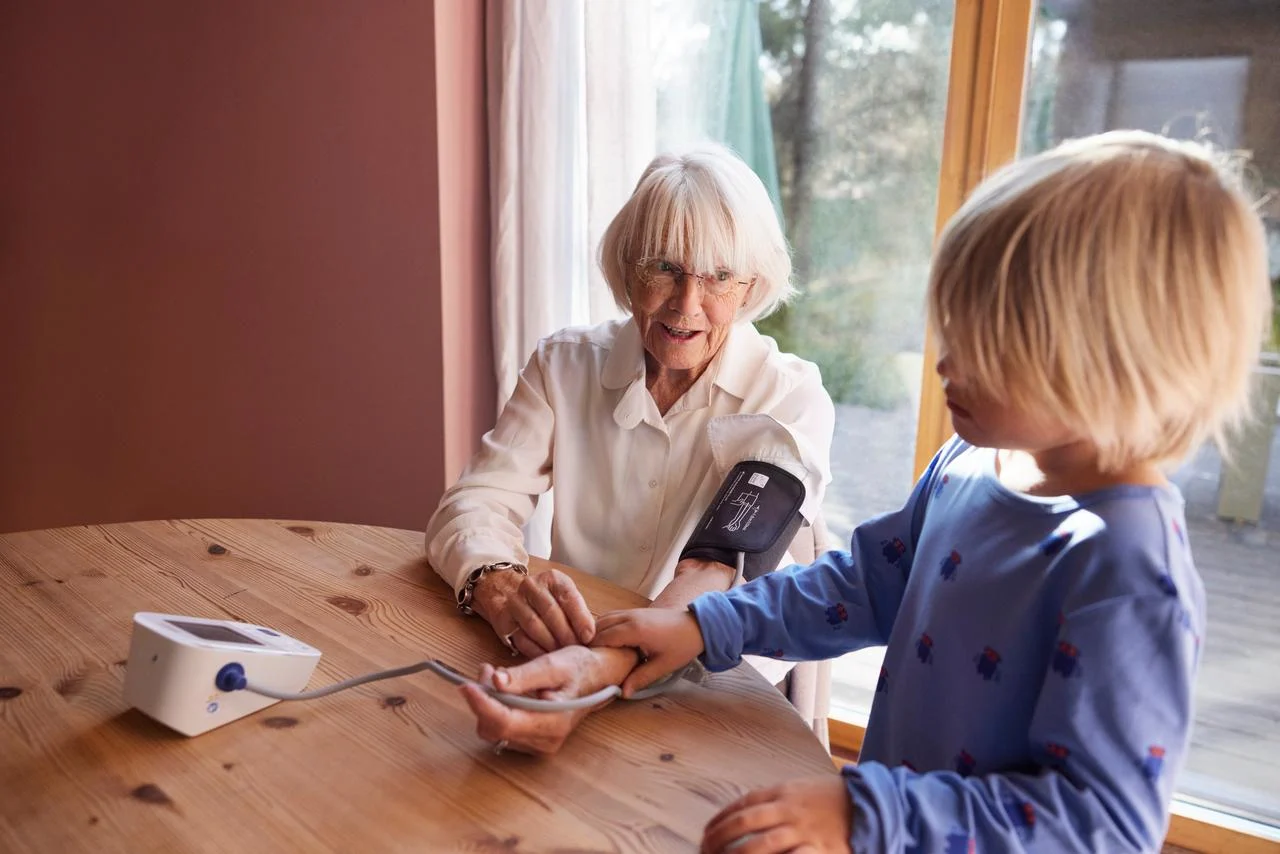 Äldre person sitter på en stol och mäter sitt blodtryck med en blodtrycksmätare. En yngre person står bredvid och känner på handleden.
