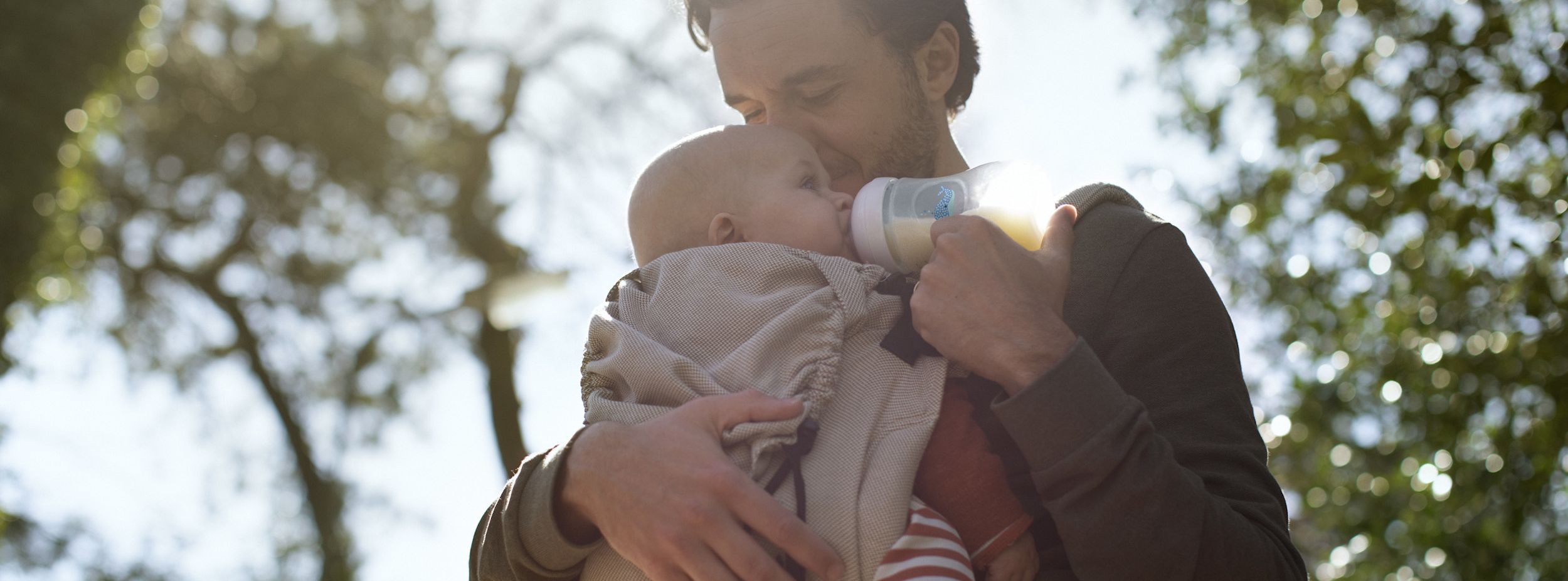 En pappa matar sitt spädbarn med nappflaska utomhus, omgiven av träd och solljus.